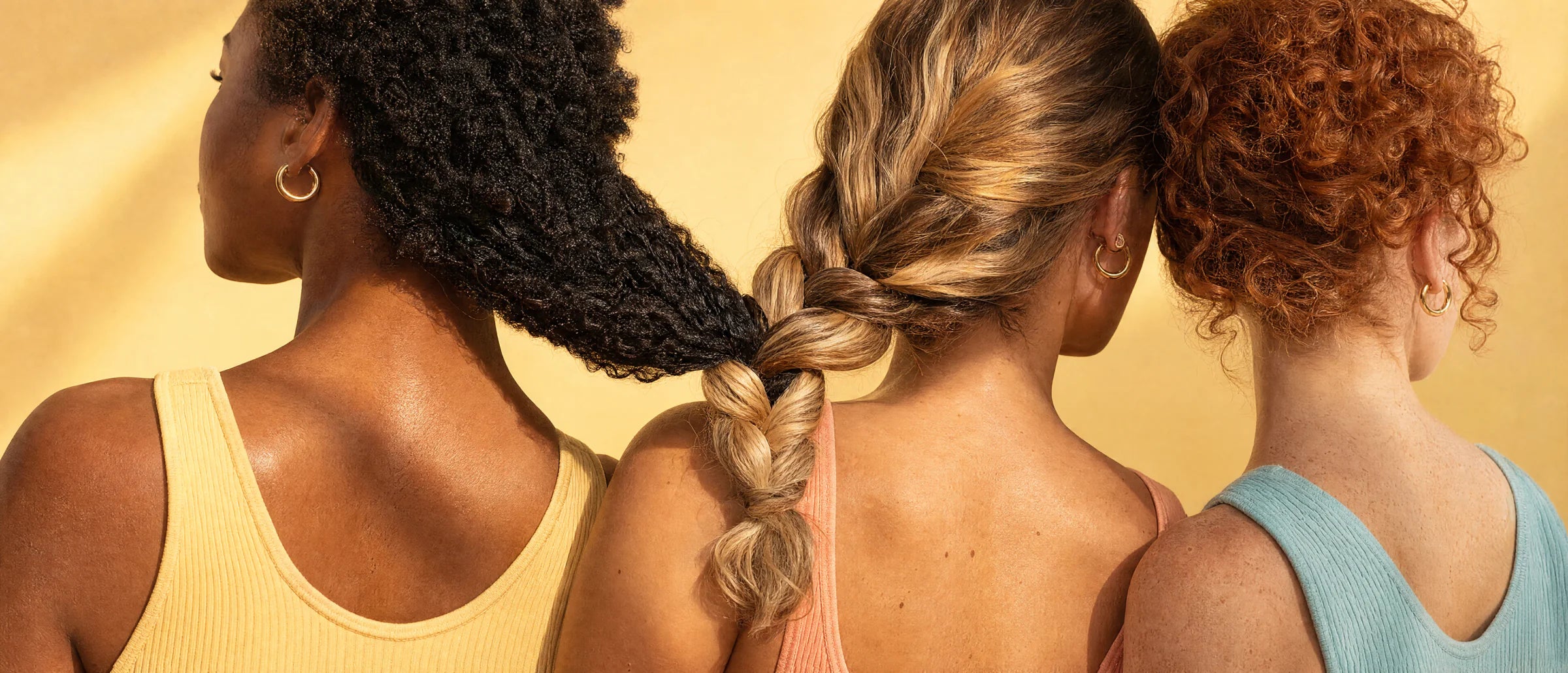 Trois femmes de dos, leurs cheveux tressés ensemble&nbsp;: afro brun, ondulés blonds, bouclés roux. Lumière dorée.